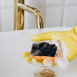 Close-up of a yellow-gloved hand cleaning with a foamy sponge under a gold faucet in a sink.